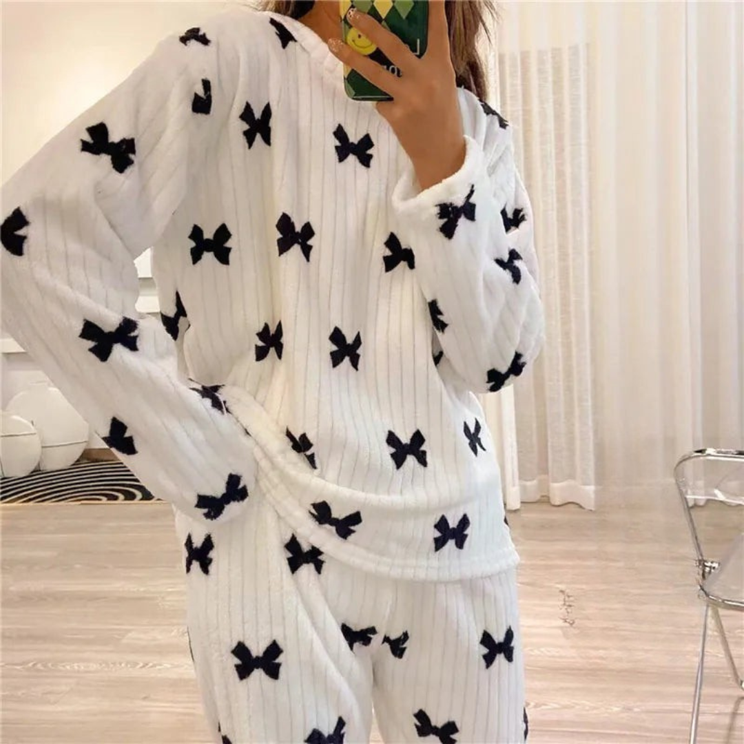 Pyjama femme en peluche blanche avec des motifs de nœuds noirs, comprenant un haut à manches longues et un pantalon élastiqué. La personne porte le pyjama tout en prenant un selfie avec un téléphone portable, offrant un look confortable et tendance pour les moments de détente à la maison.