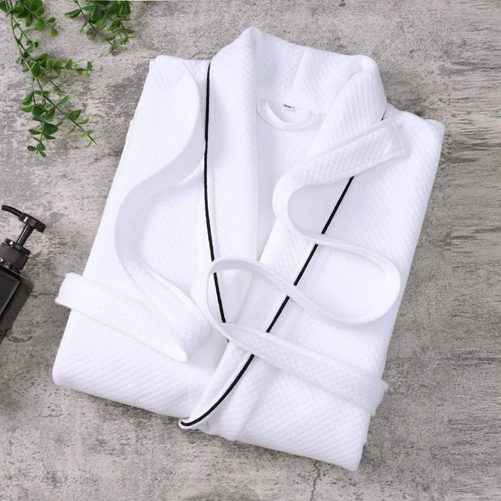 Peignoir Michel pour homme, modèle doux et agréable en tissu texturé blanc avec liseré noir, présenté plié sur une surface en pierre avec éléments de décoration.