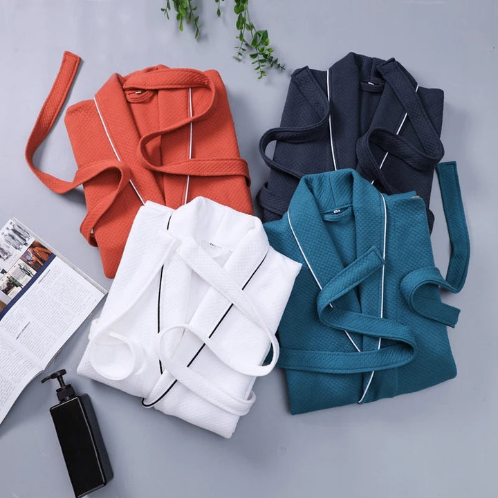 Quatre peignoirs Michel pour homme, modèles doux et agréables, présentés pliés dans différentes couleurs : orange, bleu marine, blanc et bleu pétrole, disposés sur une surface claire avec accessoires de salle de bain.