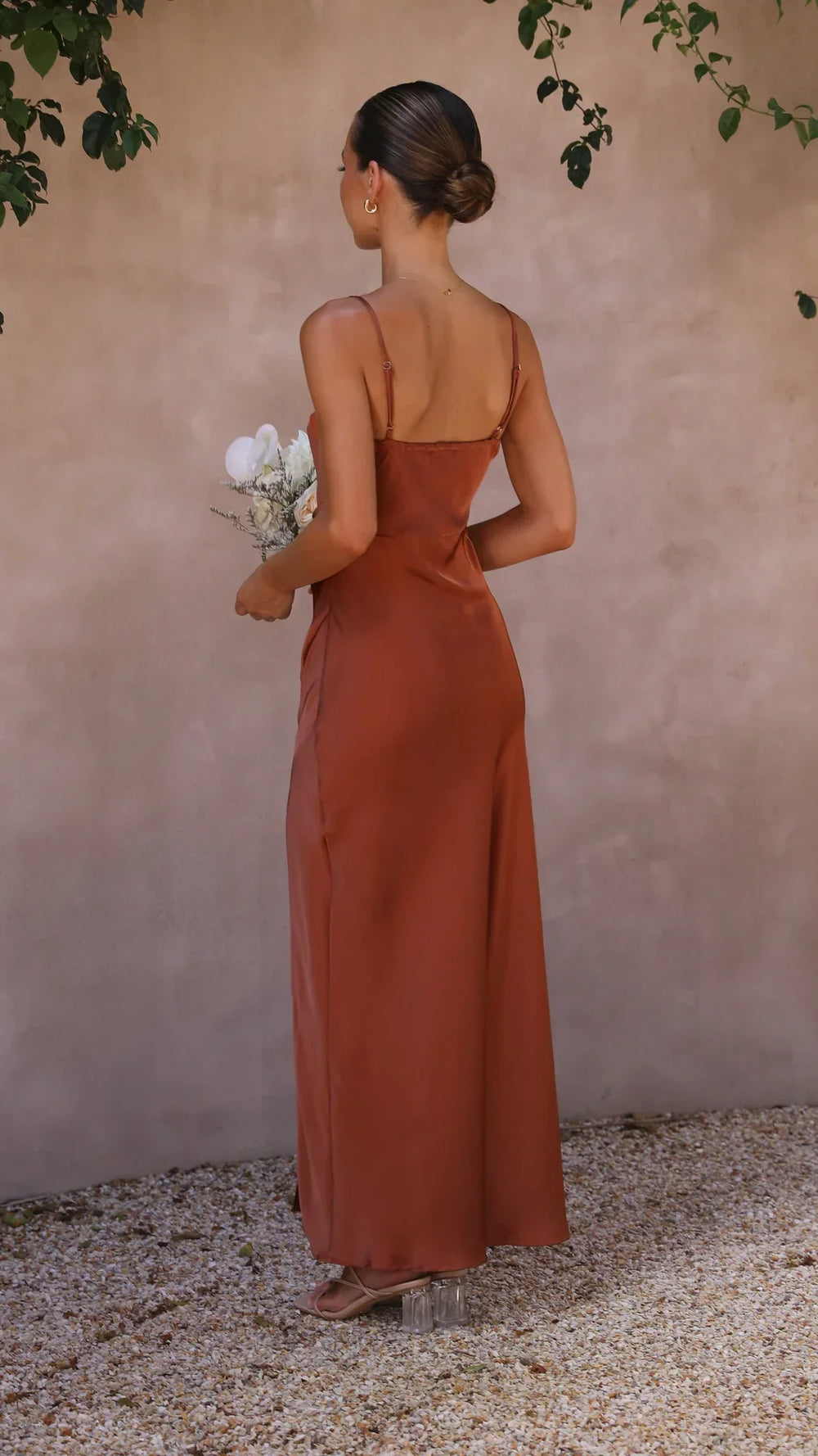 Vue arrière d'une femme portant une robe longue en satin couleur terracotta. La robe présente un design simple et élégant avec des bretelles fines. Elle tient un bouquet de fleurs blanches et sa coiffure est soignée, créant une silhouette raffinée et sophistiquée, idéale pour des occasions spéciales.