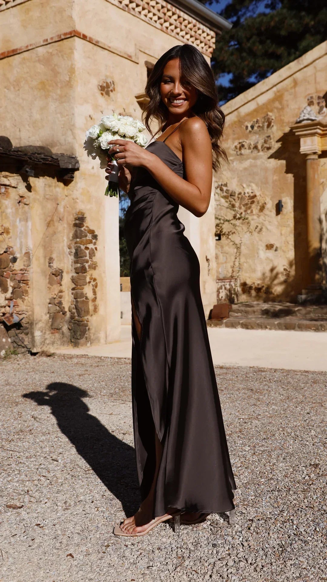 Une femme souriante porte une longue robe en satin noir fendue sur le côté. Elle tient un bouquet de fleurs blanches et porte des sandales transparentes. La coupe fluide et chic de la robe met en valeur sa silhouette, créant une allure sophistiquée, idéale pour les occasions spéciales et les soirées élégantes.