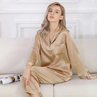 Femme portant le pyjama Mirella en satin beige, un modèle doux et confortable avec un haut à boutons et un pantalon assorti, décoré de bords satinés, assise élégamment sur un canapé moderne.