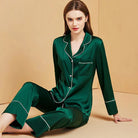 Femme portant le pyjama Mirella en satin vert, un modèle doux et confortable avec un haut à boutons et un pantalon assorti, décoré de bords blancs élégants, assise sur le sol avec un sourire détendu.