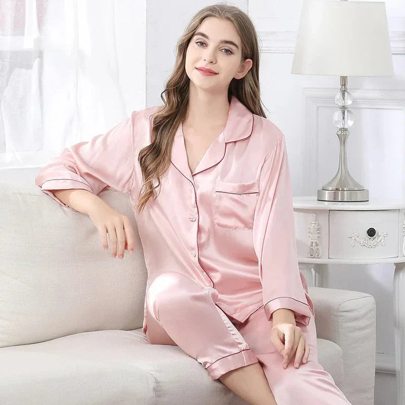 Femme portant le pyjama Mirella en satin rose, un modèle doux et confortable avec un haut à boutons et un pantalon assorti, décoré de bords noirs élégants, assise sur un canapé moderne avec une ambiance lumineuse.
