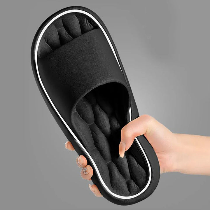 Gros plan d’un chausson noir tenu à la main, montrant la semelle intérieure matelassée et moelleuse avec large bride. Design confortable et semelle antidérapante, idéal pour un usage quotidien à la maison.