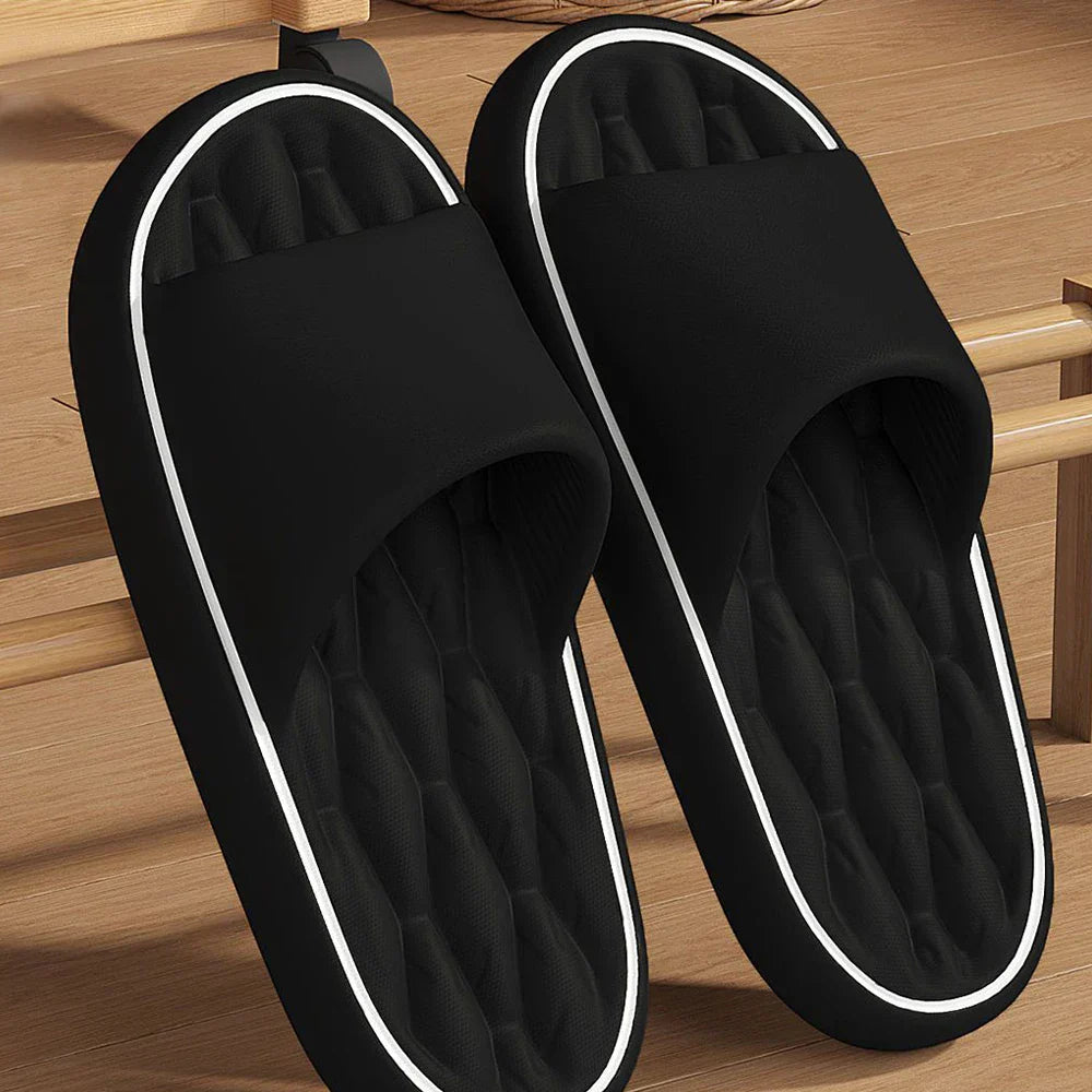 Paire de chaussons unisexes noirs posés sur un sol en bois, avec large bride et semelle intérieure matelassée pour un confort moelleux. Contours contrastés et semelle antidérapante, idéale pour la maison.