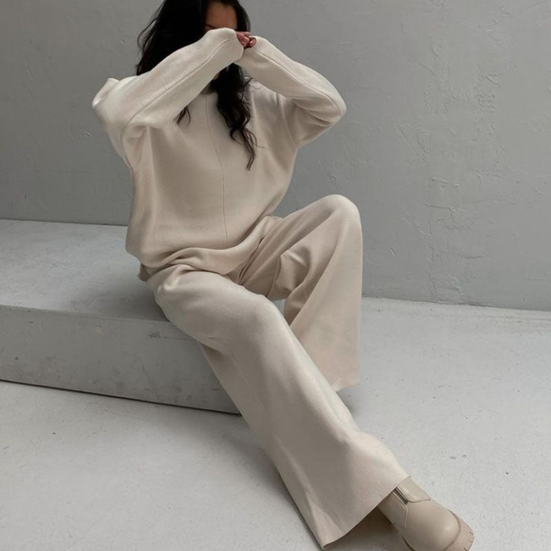Femme assise portant l’ensemble Naïla beige clair, composé d’un pull oversize à col montant et manches longues, accompagné d’un pantalon large assorti. Tenue douce, chaude et minimaliste, parfaite pour un confort optimal avec une touche élégante.