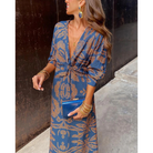 Robe portefeuille bleue à motifs dorés, décolleté en V et manches 3/4, nouée à la taille, avec bijoux dorés et pochette bleue.