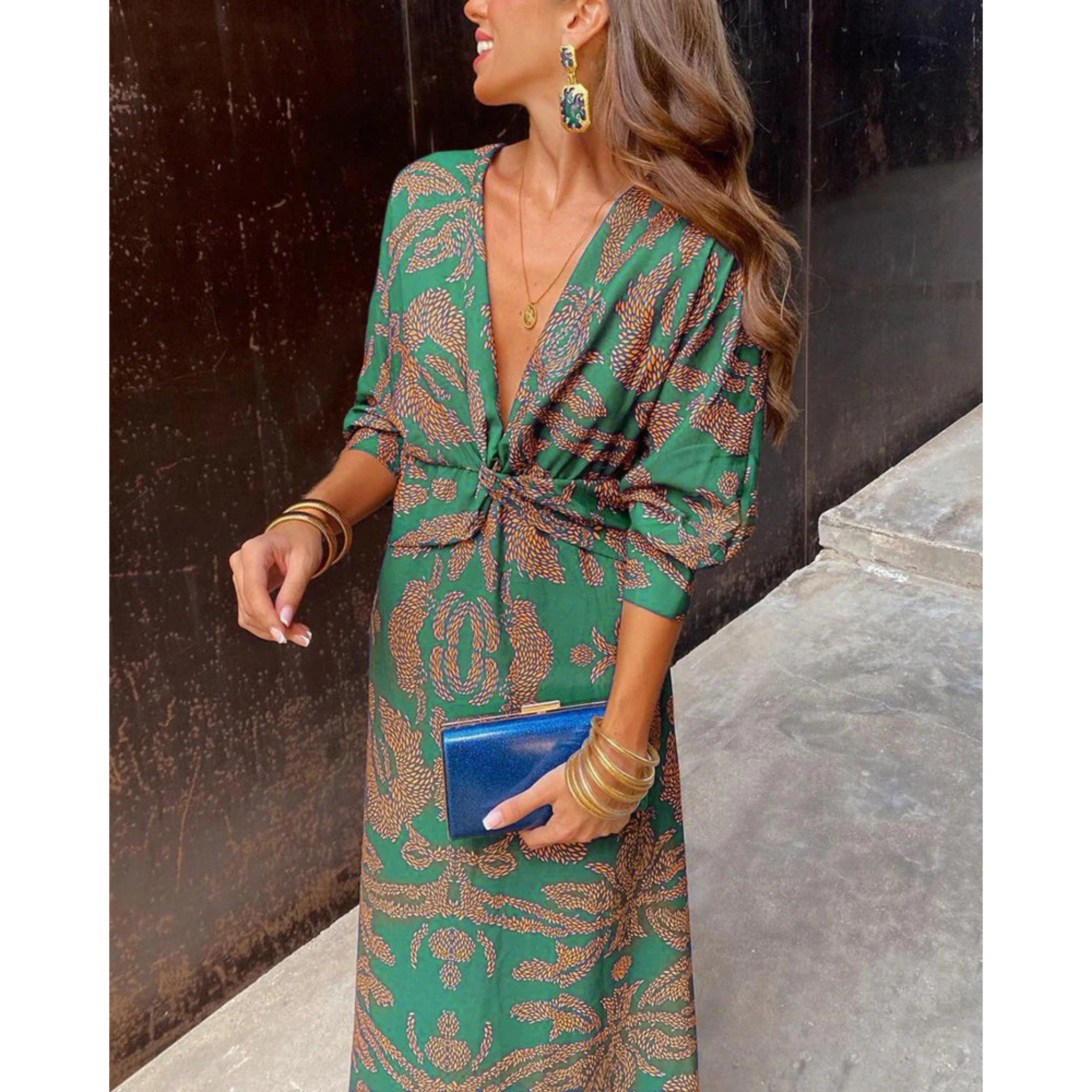 Robe portefeuille verte à motifs dorés, décolleté en V et manches 3/4, nouée à la taille, avec bijoux dorés et pochette bleue.