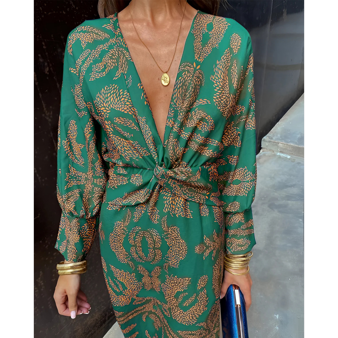 Robe portefeuille verte à motifs dorés, décolleté en V profond et taille nouée, manches longues, portée avec bijoux dorés et pochette bleue.