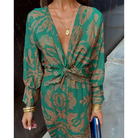 Robe portefeuille verte à motifs dorés, décolleté en V profond et taille nouée, manches longues, portée avec bijoux dorés et pochette bleue.