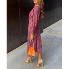 Robe midi portefeuille prune à motifs, manches longues et taille marquée, portée avec sandales à talons dorées et sac seau orange.