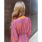 Robe prune à motifs orangés, manches longues et taille élastiquée, portée avec cheveux ondulés.