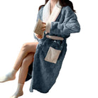 Peignoir femme chaud et confortable  Nancy en tissu peluche bleu avec col, poignets et ceinture blancs, grande poche contrastée, porté assise en intérieur avec un livre.