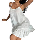 modèle en nuisette blanche fluide à fines bretelles, effet vaporeux avec volant au bas, portée assise sur un canapé.