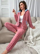 Femme assise sur un canapé beige, vêtue d’un pyjama rose en maille torsadée avec capuche doublée de bleu et poches contrastantes, offrant un style cosy et chaleureux parfait pour les moments de détente à la maison.
