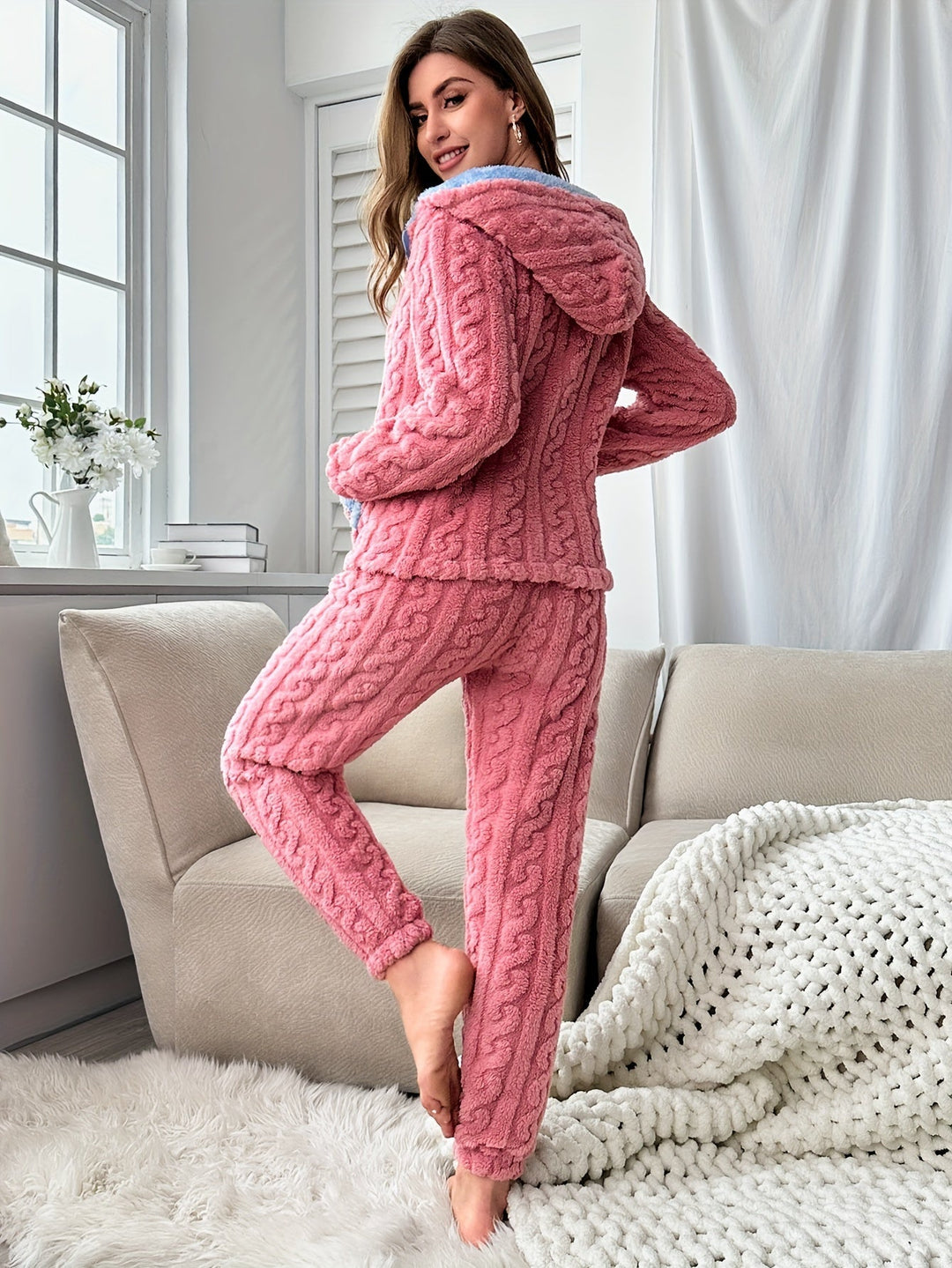 Femme debout de dos sur un tapis moelleux, vêtue d’un pyjama rose texturé avec capuche doublée de bleu, mettant en valeur la coupe ajustée et le tissu ultra doux et chaud, parfait pour les journées cocooning à la maison.
