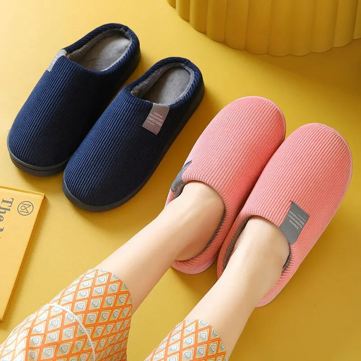 Chaussons unisexe Noé en velours côtelé, modèle rose porté aux pieds avec intérieur chaud, paire bleu marine en arrière-plan, semelles antidérapantes idéales pour la maison.