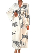Femme portant le peignoir Noelia en satin blanc avec motifs léopard noirs, coupe longue à manches larges et ceinture à nouer à la taille. Modèle tendance et agréable, idéal pour des instants de détente élégants à la maison.