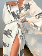 Femme portant le peignoir Noelia en satin blanc à motifs léopard noirs, noué à la taille avec une fente élégante dévoilant la jambe. Modèle fluide, tendance et agréable, parfait pour une allure sophistiquée à la maison.