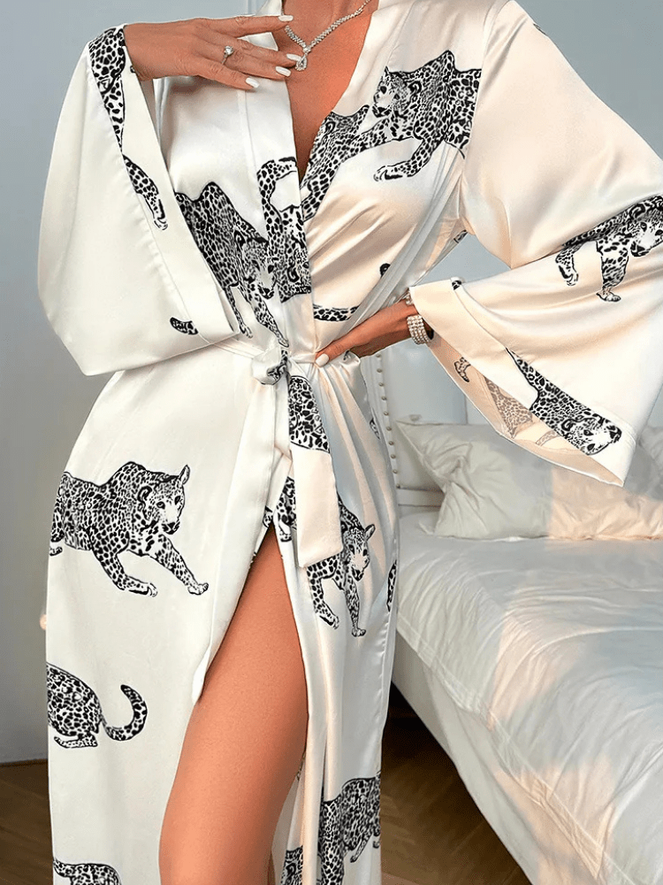 Femme portant le peignoir Noelia en satin blanc à motifs léopard noirs, noué à la taille avec une fente élégante dévoilant la jambe. Modèle fluide, tendance et agréable, parfait pour une allure sophistiquée à la maison.
