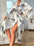 Femme assise portant le peignoir Noelia en satin blanc à imprimé léopard noir, ceinturé à la taille avec une élégante fente dévoilant la jambe. Style raffiné et tendance, idéal pour les moments de détente tout en élégance à la maison.
