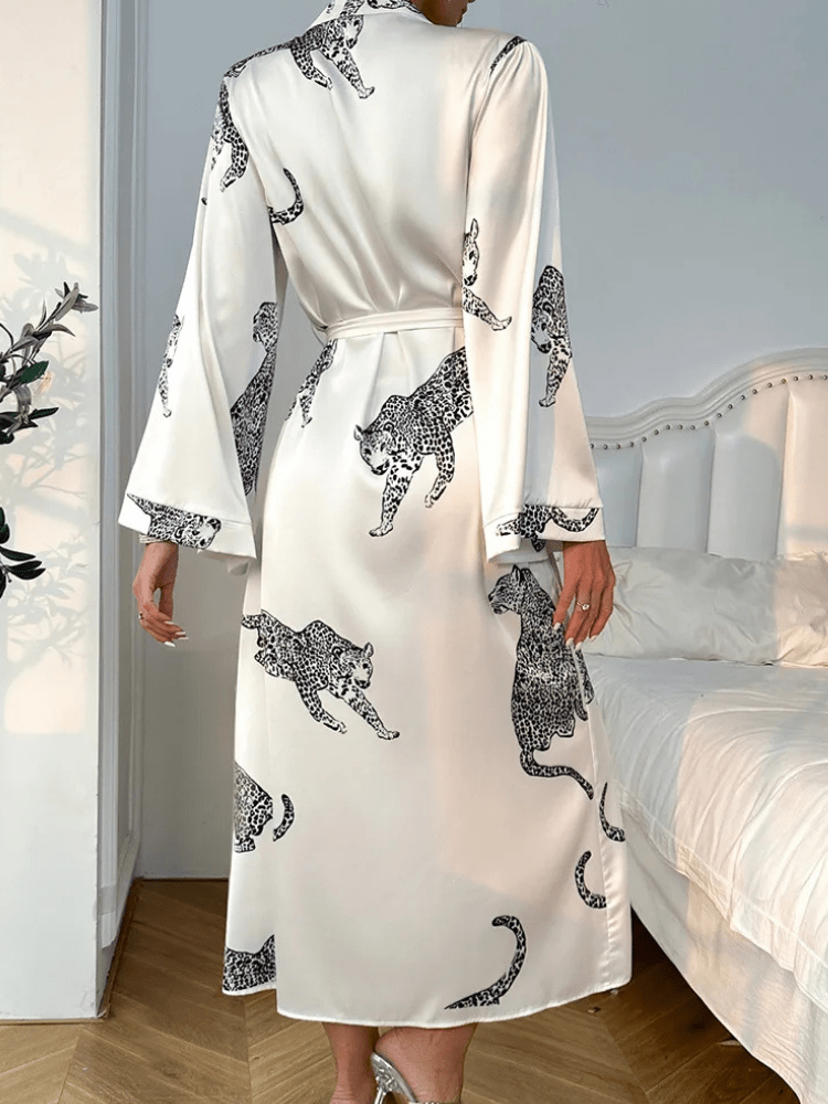 Vue arrière du peignoir Noelia porté par une femme, en satin blanc imprimé léopard noir, avec manches longues évasées et ceinture à nouer à la taille. Peignoir fluide et raffiné, parfait pour une allure élégante et confortable à la maison.
