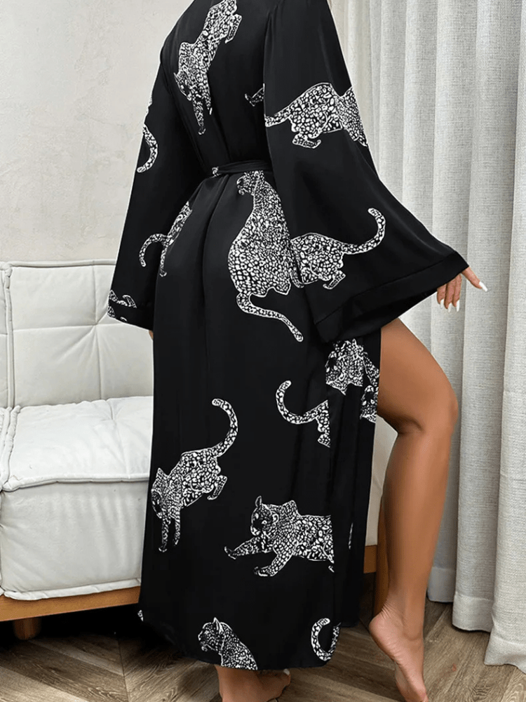 Vue arrière du peignoir Noelia en satin noir avec imprimés léopard blanc, porté par une femme révélant partiellement la jambe. Manches longues évasées et ceinture à nouer pour un look tendance, fluide et confortable à la maison.