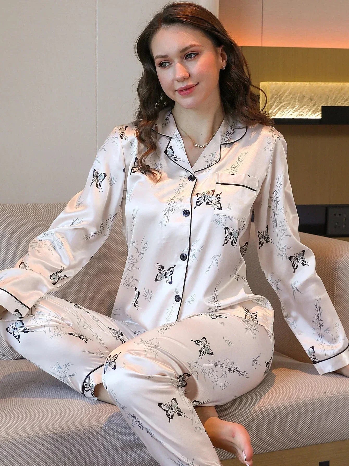 Femme assise confortablement sur un canapé, habillée d’un pyjama long satiné couleur crème à motifs papillons noirs et feuillages fins. Le haut boutonné à manches longues avec col à passepoil noir s’harmonise avec le pantalon ample. Une tenue de nuit chic et agréable, parfaite pour des moments de détente avec élégance.