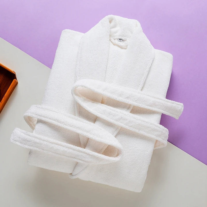Peignoir Pascal pour homme, modèle épais et chaleureux en tissu éponge blanc, soigneusement plié avec sa ceinture assortie, présenté sur un fond moderne aux tons lilas et crème.