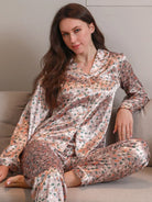Femme assise sur un canapé, portant un pyjama long en satin rose brillant avec imprimé floral coloré, composé d’un haut boutonné à manches longues et d’un pantalon assorti. Ensemble tendance et agréable à porter, parfait pour se détendre avec élégance à la maison.