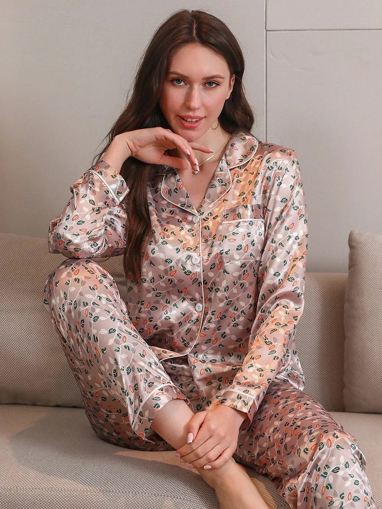 Femme assise sur un canapé, portant un pyjama long en satin rose clair à motifs floraux, composé d’un haut boutonné à col chemise et manches longues, ainsi que d’un pantalon assorti. Ensemble tendance, doux et agréable à porter, idéal pour se détendre avec élégance à la maison.