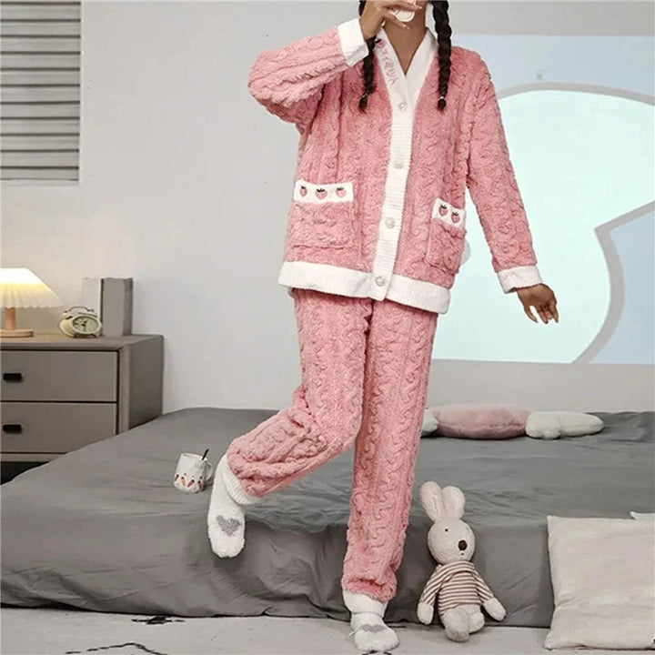 Femme portant le pyjama Percival, un ensemble d’hiver doux et chaud en maille peluche rose avec finitions blanches, composé d’un haut boutonné décoré de petits motifs et d’un pantalon assorti, idéal pour rester bien au chaud à la maison.