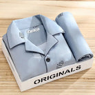 Pyjama premium pour homme présenté plié dans une boîte, en tissu bleu gris avec motifs écrits discrets. Ensemble doux, respirant et très confortable, idéal pour des nuits paisibles et élégantes.