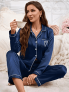 Femme assise sur un lit, tenant une tasse, vêtue d’un pyjama en satin bleu marine avec boutons blancs, passepoils contrastés et poche poitrine décorée d’un cœur stylisé. Ensemble doux, élégant et agréable à porter, idéal pour les moments de détente à la maison.