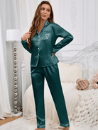 Femme debout dans une chambre élégante, portant un pyjama en satin vert émeraude, composé d’un haut à manches longues boutonné avec col à revers, passepoils blancs et poche poitrine décorée d’un cœur. Ensemble doux, brillant et agréable à porter, parfait pour des nuits chic et confortables.