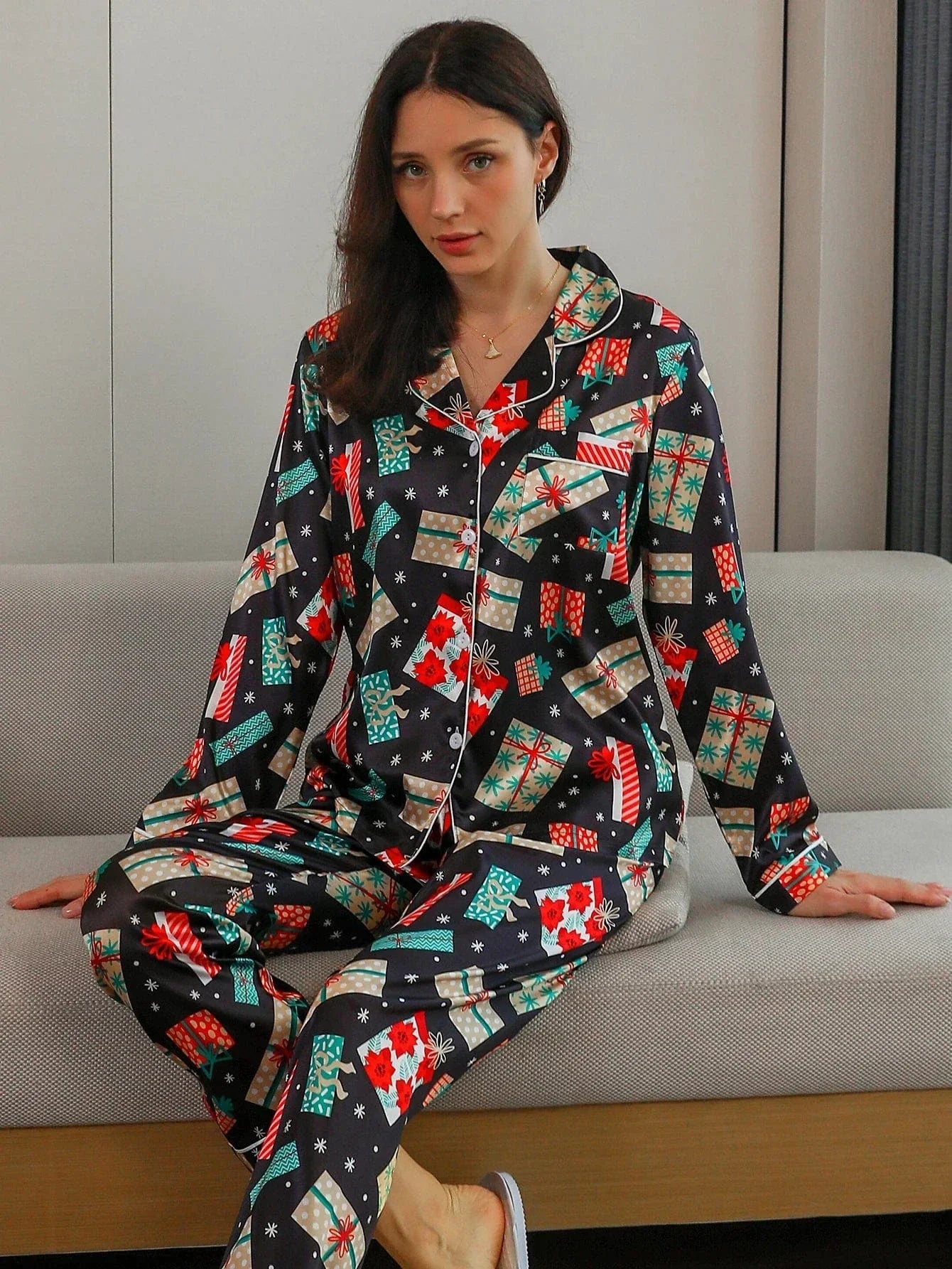 Femme assise portant le pyjama satin, un ensemble élégant et confortable spécial Noël, en satin noir décoré de motifs festifs colorés tels que cadeaux, flocons et fleurs rouges, composé d’une chemise à manches longues et d’un pantalon assorti, dans un intérieur moderne.
