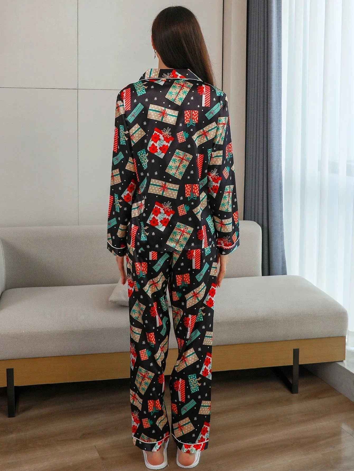 Vue de dos d’une femme portant le pyjama satin, un ensemble de Noël élégant et confortable en satin noir décoré de motifs festifs colorés cadeaux, flocons et fleurs rouges comprenant une chemise à manches longues et un pantalon assorti, photographié dans un intérieur moderne et lumineux.