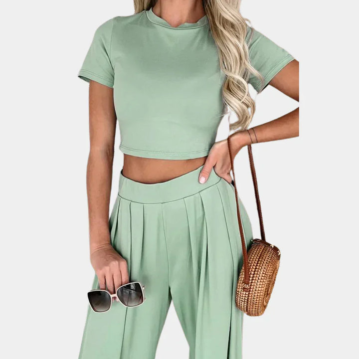 Femme portant l’ensemble Rosalind vert sauge, composé d’un crop top à manches courtes et d’un pantalon fluide taille haute à plis. Elle tient une paire de lunettes de soleil et porte un sac rond en osier. Tenue estivale légère et confortable, idéale pour un look chic et décontracté.