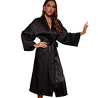 Femme dans une chambre élégante portant le peignoir Roxane en satin noir à imprimé léopard subtil. Le peignoir long et fluide est ceinturé à la taille, créant une silhouette gracieuse. L’ambiance chic de la pièce et le regard confiant de la modèle soulignent l’élégance et le raffinement de cette tenue légère parfaite pour la maison.