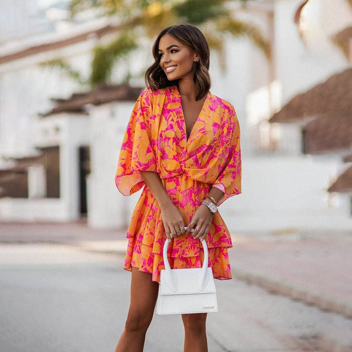 Femme souriante portant une robe mini fleurie aux couleurs vives de rose et orange, avec un design fluide et léger. Idéale pour l&