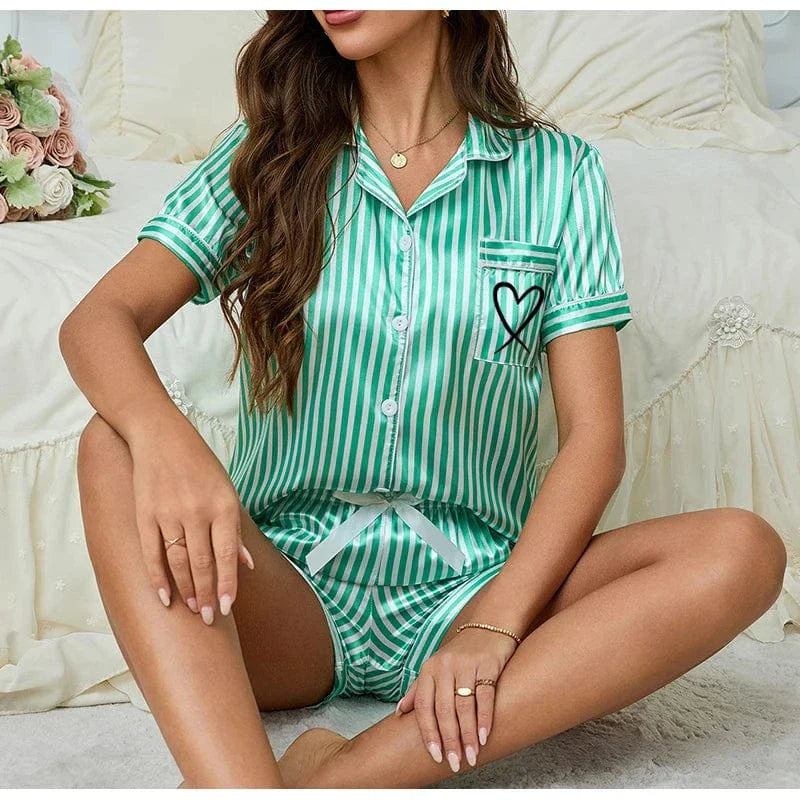 Femme assise sur le sol, portant un ensemble shorty satiné à rayures vertes et blanches. Le haut à manches courtes est boutonné, avec un col à revers et une poche poitrine décorée d’un cœur noir. Le short assorti est muni d’un cordon blanc à nouer. Tenue tendance, légère et agréable à porter, idéale pour l’été et les moments cocooning.