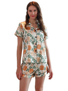Femme portant le pyjama satin, un ensemble léger, doux et élégant composé d’une chemise à manches courtes et d’un short assorti en satin blanc imprimé de fleurs orange et de feuillage vert, photographiée dans un intérieur lumineux.