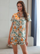 Femme portant le pyjama satin, un ensemble léger, doux et élégant en satin blanc orné de motifs floraux orange et verts, composé d’une chemise à manches courtes et d’un short assorti, photographiée dans un intérieur lumineux.