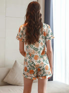 Vue de dos d’une femme portant le pyjama satin, un ensemble léger, doux et élégant en satin blanc décoré de fleurs orange et de feuillage vert, composé d’une chemise à manches courtes et d’un short assorti, photographié dans un intérieur lumineux.