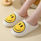 Chaussons unisexe Sasha blancs en matière pelucheuse avec grand smiley jaune, portés aux pieds, intérieur chaud et moelleux, semelle confortable pour un style cosy à la maison.