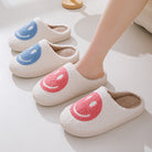 Chaussons unisexe Sasha blancs en matière pelucheuse avec smiley rose, portés aux pieds, avec modèles smiley bleu en arrière-plan, intérieur chaud et cosy pour la maison.