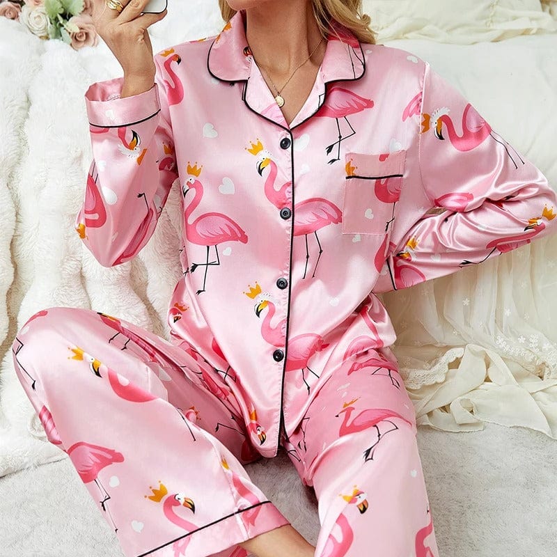 Femme assise sur un lit, vêtue d’un pyjama en satin rose imprimé de flamants roses couronnés, avec haut à manches longues boutonné, col à revers et pantalon assorti. Pyjama tendance, doux et agréable, parfait pour des nuits pleines de fantaisie et de confort.
