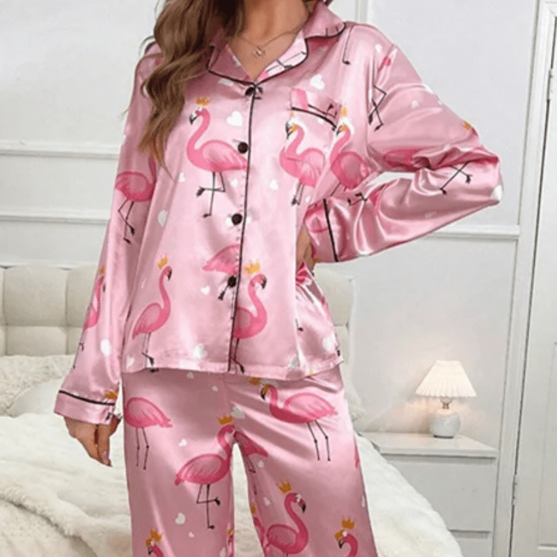 Femme portant un pyjama en satin rose à motifs flamants roses avec couronnes et petits cœurs blancs, composé d’un haut boutonné à col à revers et d’un pantalon long assorti. Pyjama tendance, ludique et confortable, parfait pour des nuits douces avec style.