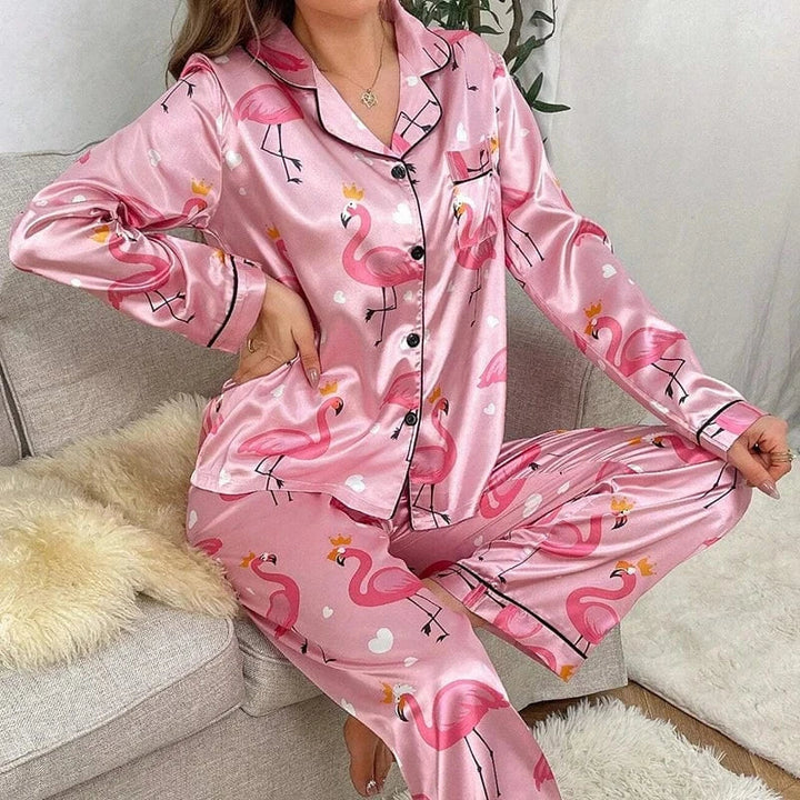 Femme assise sur un canapé, portant un pyjama en satin rose brillant à motifs flamants roses couronnés et petits cœurs blancs, composé d’un haut boutonné à col à revers et d’un pantalon long assorti. Ensemble de nuit tendance et confortable, parfait pour une ambiance cocooning avec style.